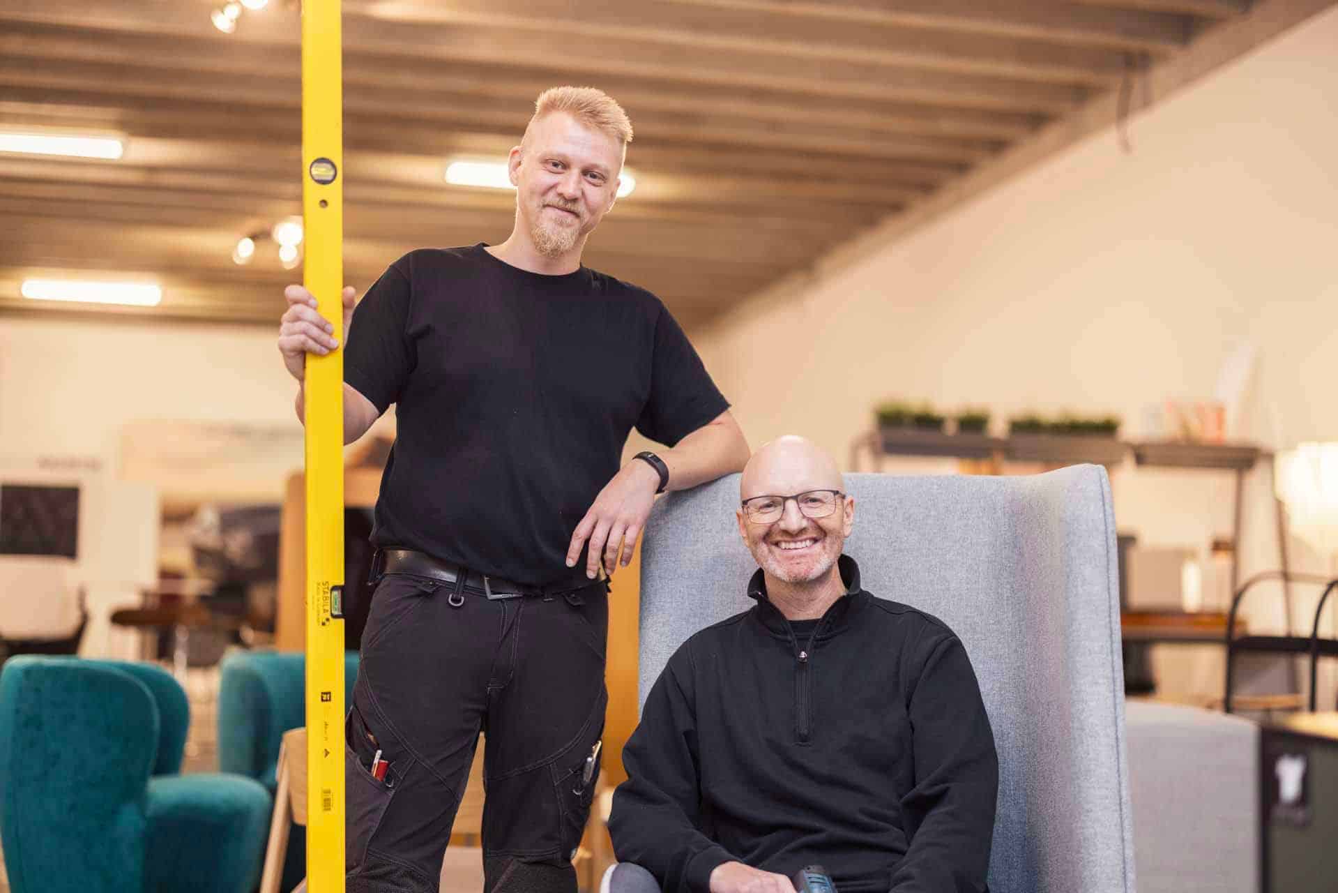 To montører i et showroom for kontormøbler, den ene står med et gult vaterpas og den anden sidder i en grå lænestol
